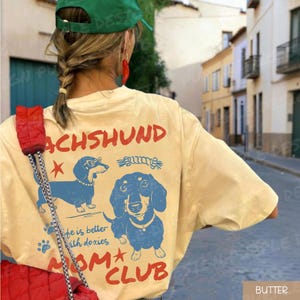 May include: A light yellow t-shirt with a dachshund graphic. The shirt features the word "DACHSHUND" in red, along with two blue dachshund illustrations and the text "life is better with doxies MOM CLUB".