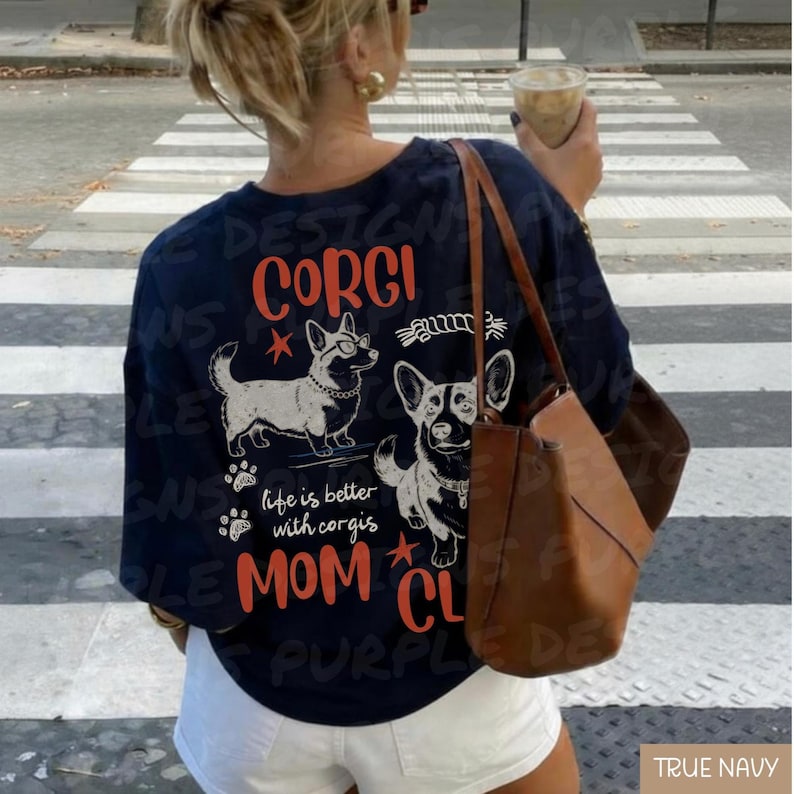 Puede incluir: Camiseta azul marino con un dise&ntilde;o gr&aacute;fico que presenta dos corgis y las palabras "CORGI MOM CLUB". El dise&ntilde;o incluye huellas de patas y la frase "life is better with corgis" en blanco y naranja.