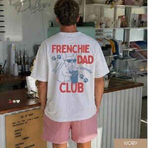 May include: White t-shirt featuring a French Bulldog graphic wearing sunglasses and drinking from a cup, with the words "FRENCHIE DAD CLUB" in red. The shirt also has blue paw prints. The person is wearing pink and white striped shorts.