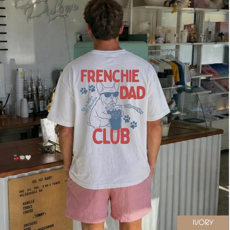 May include: White t-shirt featuring a French Bulldog graphic wearing sunglasses and drinking from a cup, with the words "FRENCHIE DAD CLUB" in red. The shirt also has blue paw prints. The person is wearing pink and white striped shorts.