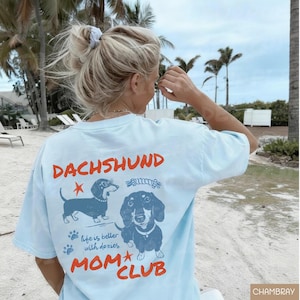 May include: Light blue t-shirt with the words "DACHSHUND MOM CLUB" in red and a graphic of two dachshunds. The shirt also has the phrase "life is better with doxies" in a script font.