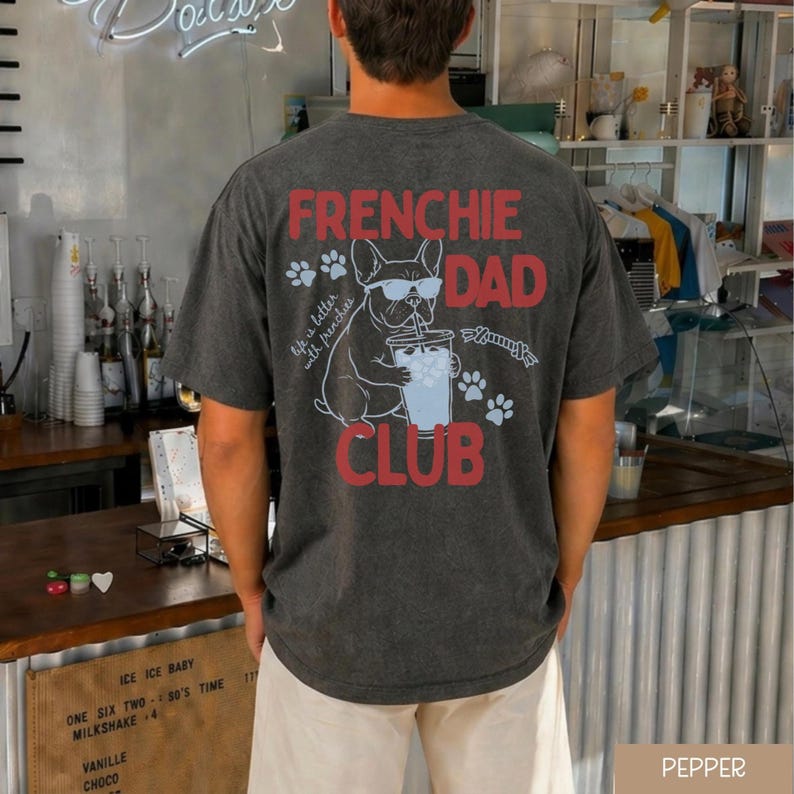 May include: Charcoal grey t-shirt with the words "FRENCHIE DAD CLUB" in red. The design includes a French Bulldog wearing sunglasses and drinking from a cup, plus paw prints. The text "life is better with frenchies" is also present.