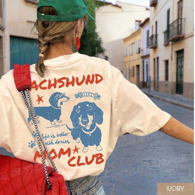 May include: Ivory-colored t-shirt with a dachshund graphic in blue and the text "DACHSHUND MOM CLUB." The shirt features two dachshund illustrations, one wearing glasses. The phrase "life is better with doxies" is also printed on the shirt.