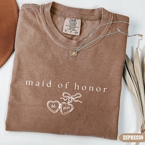 May include: A brown t-shirt with the text "maid of honor" and a small illustration of a locket with the text "Est. 2023".
