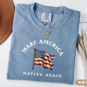 May include: Blue jean-colored t-shirt with the text "MAKE AMERICA NATIVE AGAIN" and an American flag graphic. A gold necklace with a pendant rests on the shirt. The shirt is folded and has a Comfort Colors tag.
