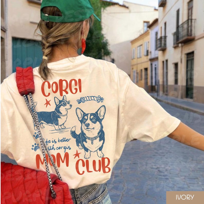 Puede incluir: Camiseta de color marfil con dos ilustraciones de perros corgi en azul y negro, con las palabras "CORGI MOM CLUB" en rojo. La camiseta tambi&eacute;n incluye la frase "life is better with corgis" en azul.