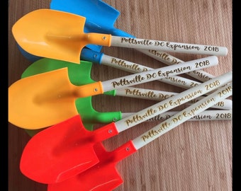 May include: Five colorful plastic toy shovels with wooden handles. The handles are decorated with gold lettering that reads "Pottsville DC Expansion 2018".
