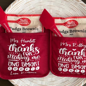 May include: Two red oven mitts with pockets. Each mitt has a Betty Crocker Fudge Brownie mix box inside. The mitts have white text that says "Mrs. Hamlett thanks for making me one smart cookie Love Kadence" and "Mrs. Riffey thanks for making me one smart cookie Love Kate".