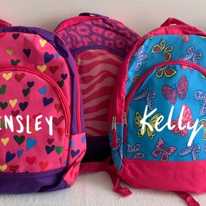 May include: Five colorful backpacks with various patterns, including hearts, butterflies, flowers, and owls. The backpacks have names like "Kinsley," "Kelly," and "Mia" printed on them. Each backpack has a different design and color scheme.