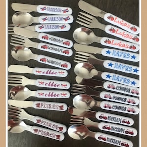 May include: A set of 12 personalized children's cutlery sets with various designs. The handles are white with colorful designs and names printed on them. The designs include airplanes, fire trucks, stars, unicorns, bears, and tractors.