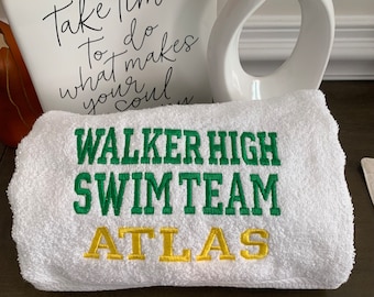 May include: White towel embroidered with "WALKER HIGH SWIM TEAM ATLAS" in green and gold lettering. The towel is folded and rests on a dark surface. A white decorative vase and a ceramic piece with script are in the background.