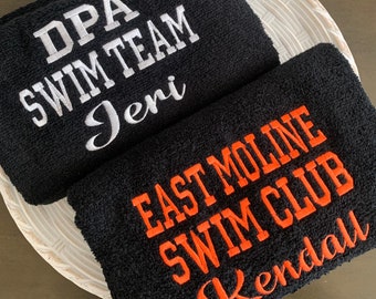 May include: Two black embroidered towels. One towel has "DPA SWIM TEAM" in white block letters and "Jeri" in cursive. The other towel has "EAST MOLINE SWIM CLUB" in orange block letters and "Kendall" in cursive.