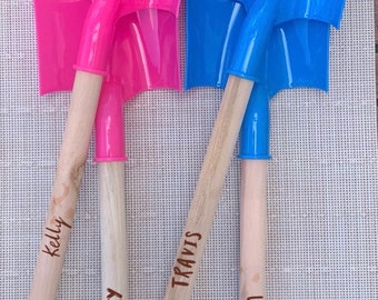 May include: Three wooden handled shovels with personalized names. The shovels are pink, blue, and blue. The names are "Kelly", "Aubrey", and "Jaxson".