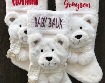 May include: Three white plush Christmas stockings with bear faces and scarves. Each stocking has a name embroidered on the cuff: "Giovanni" in red, "Grayson" in red, and "Baby Bialik" in purple.