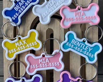 May include: A collection of colorful, bone-shaped pet ID tags with silver key rings. Each tag features a different name and phone number in white text against backgrounds of blue, white, yellow, pink, teal, purple, and lavender.