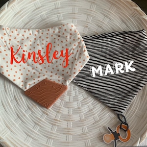 May include: Two baby bibs with personalized names. One bib is white with brown polka dots and the name "Kingsley" in orange text. The other bib is black with white stripes and the name "Mark" in white text. Both bibs have a brown silicone teether attached.