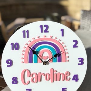 May include: A white, round wall clock with a rainbow design in pink, purple, and teal. The clock features the name "Caroline" in pink script and purple numerals. The clock hands are black.