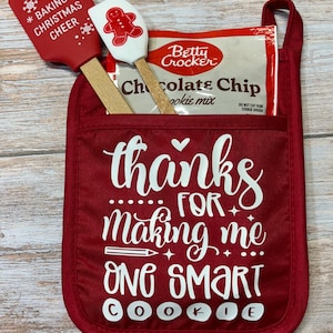 May include: Red oven mitt with a pocket containing a box of chocolate chip cookie mix and two red and white spatulas. The oven mitt has the text "thanks for making me one smart cookie" in white.