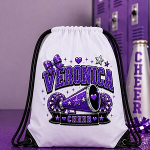 May include: White drawstring bag with a purple glitter design. The design includes the name "VERONICA" in large purple letters, a bow, stars, hearts, and a megaphone. The word "CHEER" is on a black banner.