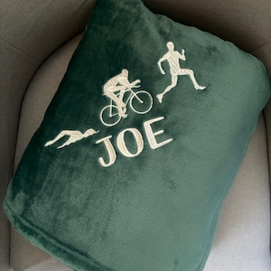 Könnte beinhalten: Ein dunkelgrünes Plüschkissen mit weißer Stickerei einer Person auf dem Fahrrad, einer Person, die rennt, und dem Namen "JOE".