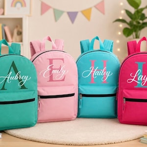 May include: Four personalized backpacks in teal, pink, and turquoise. Each backpack features a name in a cursive font with a large initial. The names are Aubrey, Emily, Hailey, and Layla. The backpacks have black zippers and straps.