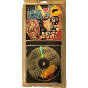 May include: A sealed Jukebox Giants Volume 3 CD featuring WCBS FM 101 branding. The cover art displays a cartoon figure, a jukebox, and the text Jukebox Giants Volume 3. The disc is visible in the lower section of the clear plastic packaging.