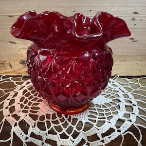 May include: A ruby red glass bowl with a ruffled rim and diamond pattern. The bowl sits on a white crocheted doily. The bowl has a round base and a textured surface. The background is a wooden surface.