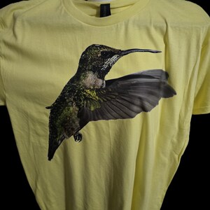 Puede incluir: Camiseta amarillo claro con un gráfico detallado de un colibrí en vuelo. El ave tiene plumaje verde, marrón y negro con alas grises extendidas. La camiseta es de material suave y tiene cuello redondo.