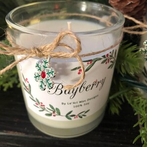 Puede incluir: Una vela de vidrio transparente con un interior de cera blanca y una etiqueta decorativa que dice "Bayberry". La etiqueta presenta un dise&ntilde;o floral rojo y verde. La vela est&aacute; atada con hilo y tiene un dije de copo de nieve.