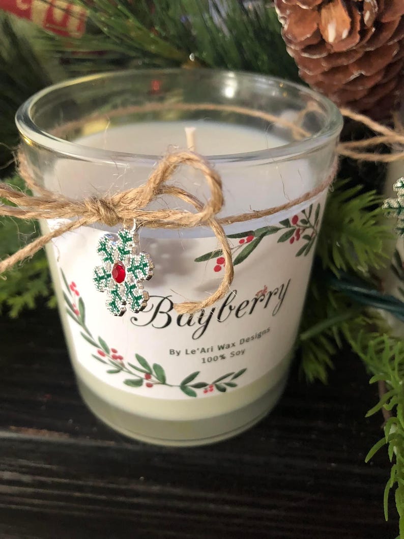 Puede incluir: Una vela de vidrio transparente con un interior de cera blanca y una etiqueta decorativa que dice "Bayberry". La etiqueta presenta un dise&ntilde;o floral rojo y verde. La vela est&aacute; atada con hilo y tiene un dije de copo de nieve.
