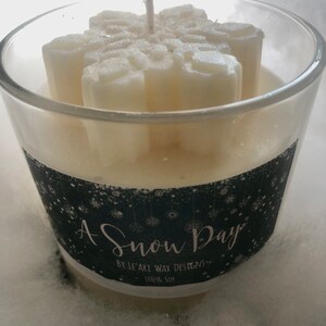 Puede incluir: Un portavelas de cristal transparente con una vela en forma de copo de nieve en su interior. La vela es blanca y tiene una mecha en el centro. La etiqueta dice "A Snow Day" y "100% Soy".