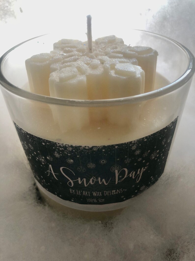 Puede incluir: Un portavelas de cristal transparente con una vela en forma de copo de nieve en su interior. La vela es blanca y tiene una mecha en el centro. La etiqueta dice "A Snow Day" y "100% Soy".