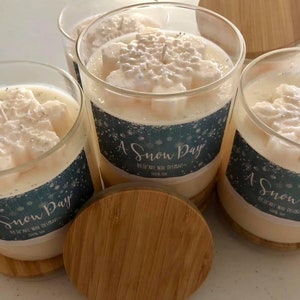 Puede incluir: Cuatro velas de vidrio con tapas de madera. Las velas tienen un dise&ntilde;o de copo de nieve en la parte superior y una etiqueta que dice "A Snow Day". Las velas est&aacute;n hechas de cera cremosa y 100% soja.