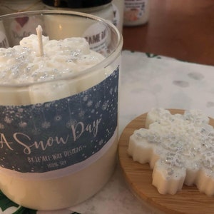 Puede incluir: Un portavelas de cristal transparente con una vela en forma de copo de nieve en su interior. La vela es blanca con detalles de purpurina plateada. Un derrite de cera con forma de copo de nieve a juego se encuentra sobre un peque&ntilde;o posavasos de madera. La etiqueta dice "A Snow Day".