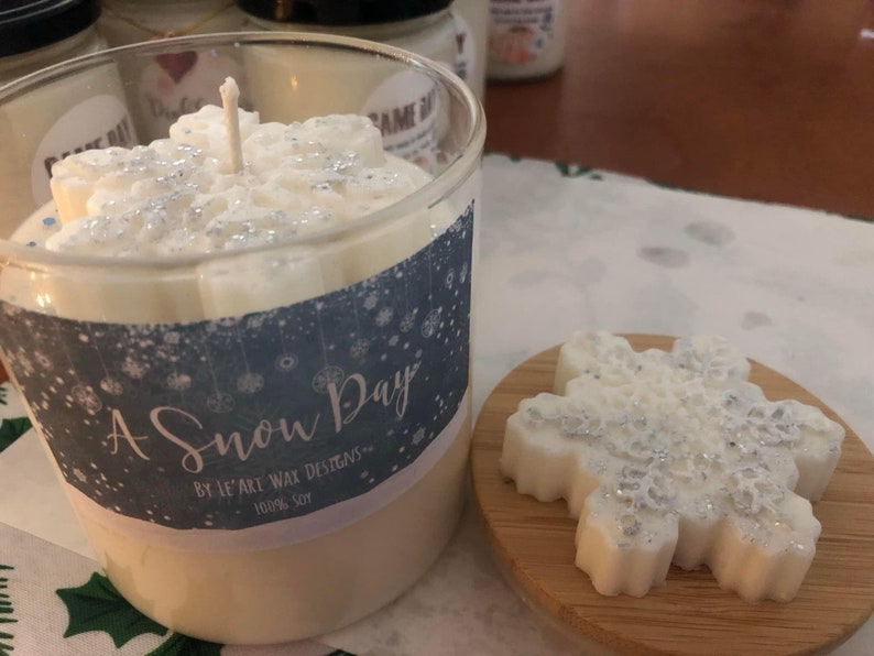 Puede incluir: Un portavelas de cristal transparente con una vela en forma de copo de nieve en su interior. La vela es blanca con detalles de purpurina plateada. Un derrite de cera con forma de copo de nieve a juego se encuentra sobre un peque&ntilde;o posavasos de madera. La etiqueta dice "A Snow Day".