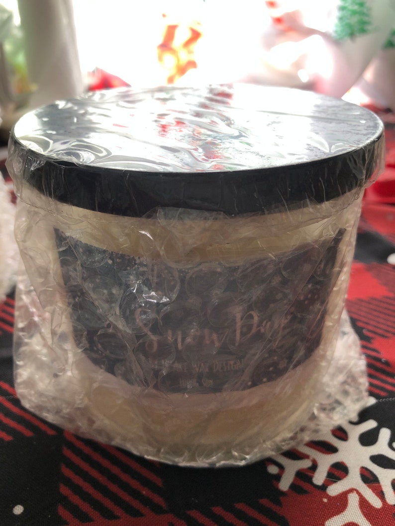 Puede incluir: Una vela redonda en un recipiente blanco con tapa negra, envuelta en pl&aacute;stico de burbujas. La etiqueta dice "Snow Day" en escritura blanca. La vela est&aacute; sobre una superficie a cuadros rojos y negros.