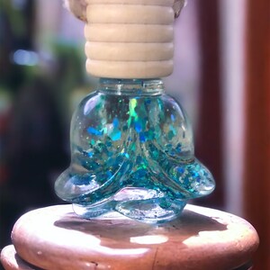 May include: A clear, bell-shaped car air freshener filled with blue and green glitter. It has a light-colored wooden top and a rope for hanging. The air freshener sits on a wooden surface.