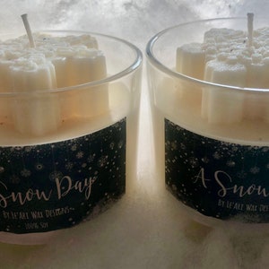 Puede incluir: Dos portavelas de cristal transparente con velas en forma de copo de nieve. Las velas son blancas y tienen una mecha. Los portavelas tienen una etiqueta negra con el texto "A Snow Day" y "100% Soy".
