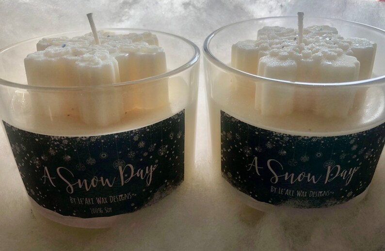 Puede incluir: Dos portavelas de cristal transparente con velas en forma de copo de nieve. Las velas son blancas y tienen una mecha. Los portavelas tienen una etiqueta negra con el texto "A Snow Day" y "100% Soy".