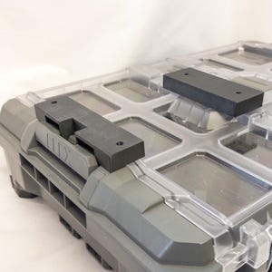 May include: A gray and clear plastic storage container with a clear lid. The container has several compartments and black latches. The lid has black handles and is secured with black clips. The container is designed for tool storage.