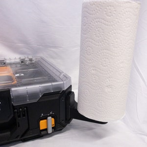 May include: A black and orange tool organiser with a clear lid, holding a roll of white paper towels. The paper towel holder is attached to the side of the organiser. The paper towel roll is white with a repeating pattern.