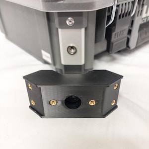 Puede incluir: Un objeto negro, angular, impreso en 3D con un agujero central y sujetadores dorados. El objeto está posicionado frente a un contenedor de almacenamiento gris con una bisagra plateada y un asa negra.