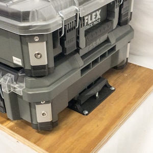 以下が含まれることがあります： グレーのFLEX STACK PACKツールボックスが2つ積み重ねられています。上のボックスは透明な蓋と金属製の留め金が付いています。ボックスは、黒い金属製のブラケットが付いた木製のプラットフォームに置かれています。