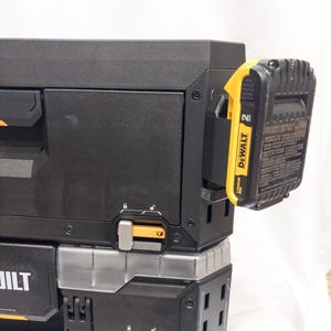 Puede incluir: Sistema de almacenamiento de herramientas negro y amarillo con una batería adjunta. El sistema tiene múltiples compartimentos y mecanismos de bloqueo. La batería está etiquetada como "DEWALT" y "2 Ah".