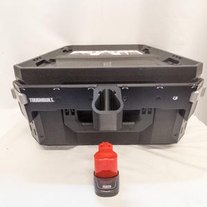 Könnte beinhalten: Schwarze Toughbuilt-Werkzeugbox mit einem schwarzen und roten Milwaukee M12 Lithium-Ionen-Akku. Die Werkzeugbox hat einen Griff und steht auf einer weißen Oberfläche. Das Toughbuilt-Logo ist auf der Vorderseite sichtbar.