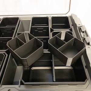 Puede incluir: Una caja de almacenamiento de plástico gris con una tapa transparente, que presenta múltiples insertos de compartimentos negros. Los insertos son de varias formas y tamaños, diseñados para organizar objetos pequeños. La caja tiene un cierre seguro.