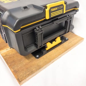 Op de afbeelding: Zwarte en gele ToughSystem gereedschapskist met een handvat en sluitingen. De gereedschapskist is gemonteerd op een houten plank. De tekst "TOUGHSYSTEM" is zichtbaar. Ontworpen voor het opbergen van gereedschap.