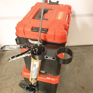 Könnte beinhalten: Eine rote und schwarze Craftsman-Werkzeugkiste mit einer schwarzen Kartuschenpistole und einem schwarzen Becherhalter. Die Kartuschenpistole enthält eine Tube DAP-Dichtmasse. Die Werkzeugkiste ist auf einer weiteren schwarzen Werkzeugkiste gestapelt.