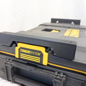 Op de afbeelding: Zwarte en gele ToughSystem gereedschapsopbergbox met een zwart deksel en gele accenten. De box heeft een gele sluiting met het ToughSystem logo. De box is ontworpen voor duurzaamheid en organisatie.