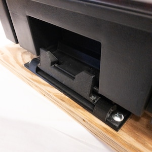 Może przedstawiać: Zbliżenie czarnego plastikowego elementu z prostokątnym otworem i mechanizmem przesuwnym. Element jest zamontowany na drewnianej powierzchni i zabezpieczony śrubą. Projekt wydaje się funkcjonalny i przemysłowy.